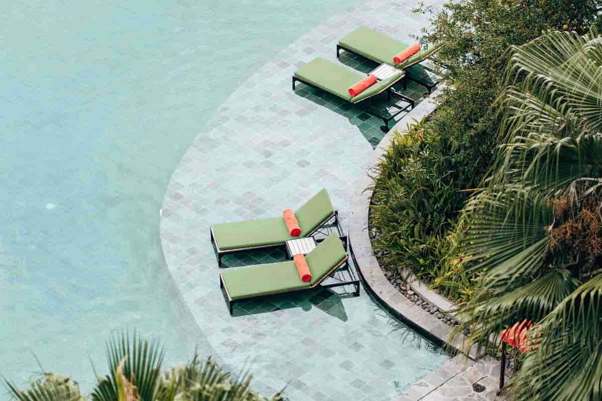 A pool seen from above with two green lounge chairs on a tiled shelf beside palm leaves