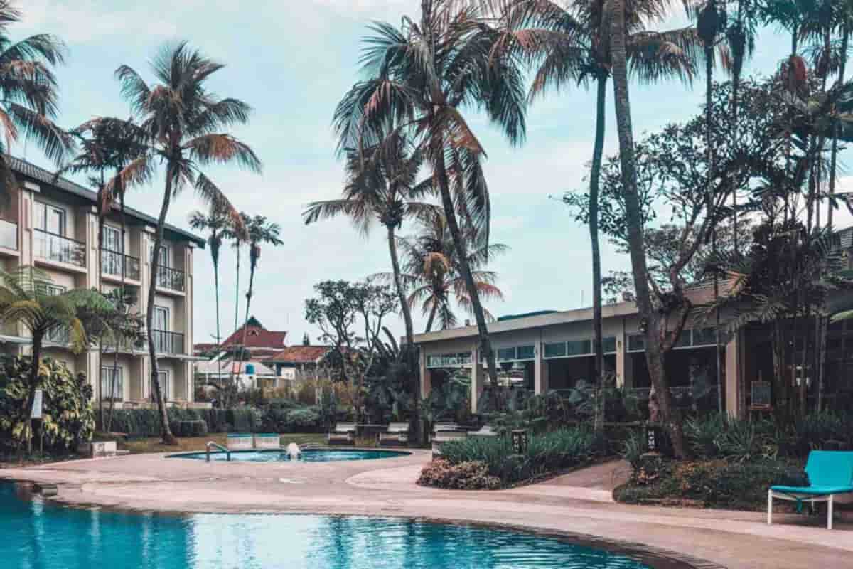 A curved pool in front of a low building and tall palm trees under a partly cloudy sky
