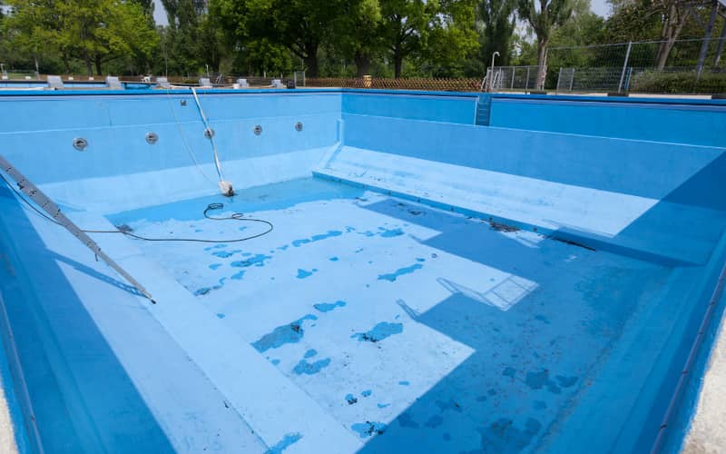 An empty pool with blue walls and floor, worn patches, a hose and a ladder lying inside