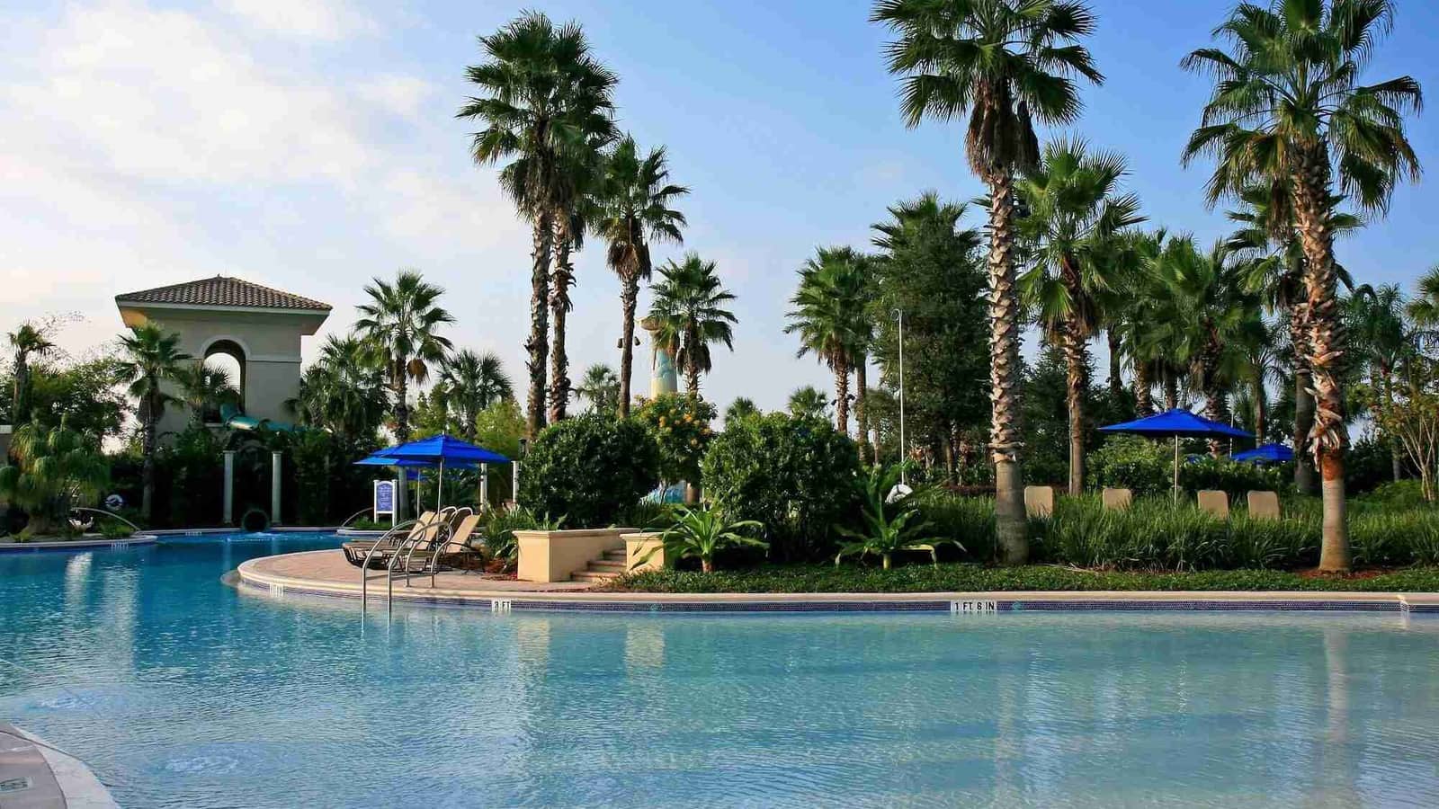 A swimming pool with a curved edge, palm trees, a pavilion and blue umbrellas under a blue sky