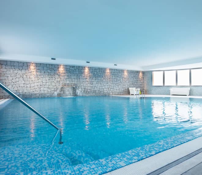 An indoor pool with a stainless handrail, a stone wall and tall windows