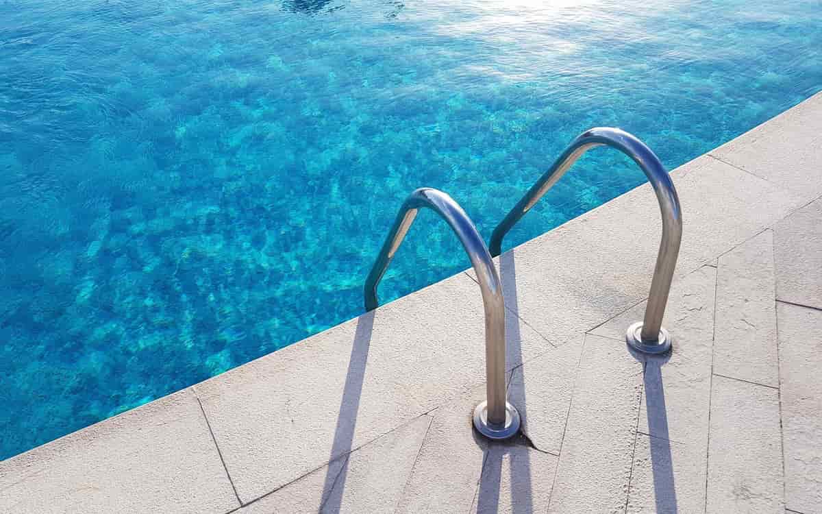 Residential Pool Repairs: a chrome pool ladder on a pale stone edge above bright blue water