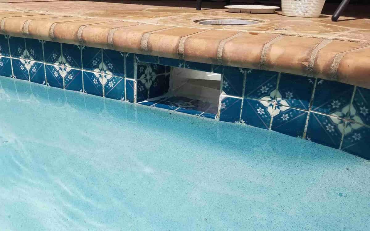 The waterline of a pool with decorated blue tiles, a skimmer opening in the wall and a stone coping above