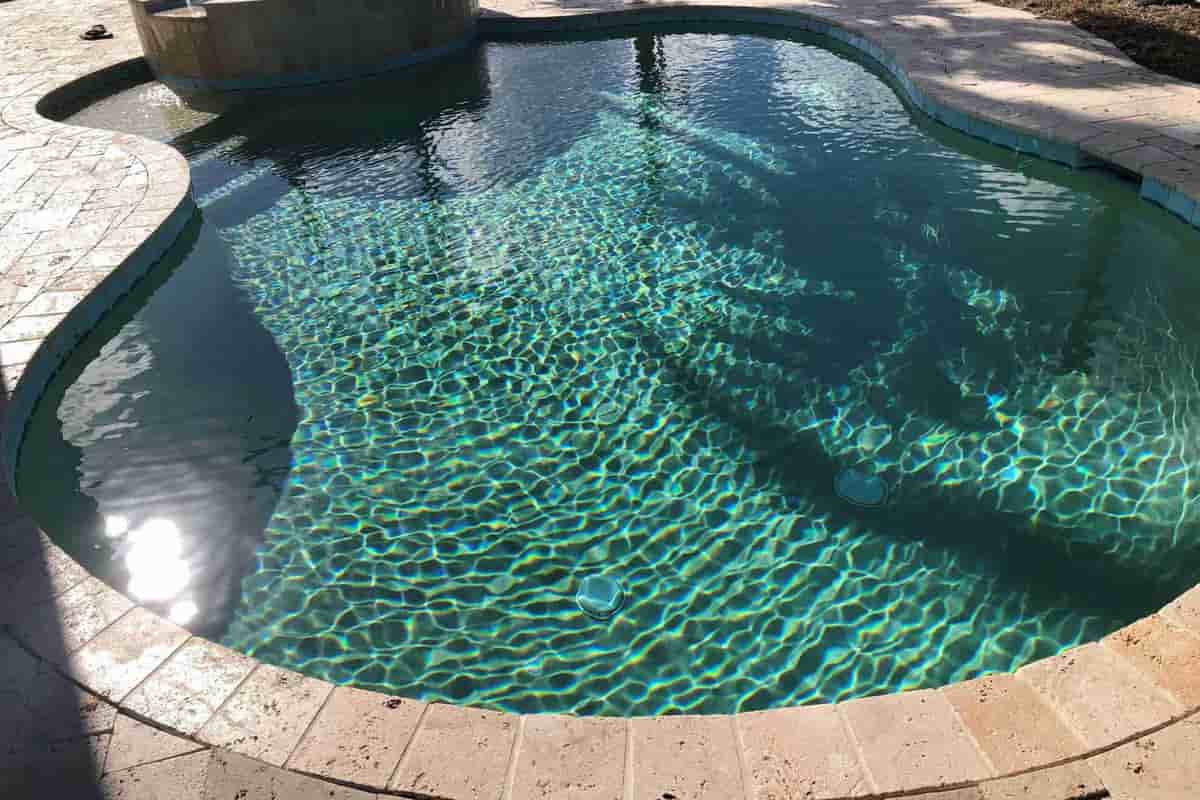 A pool with a raised spa at one end, clear rippling water and stone paving around the edge