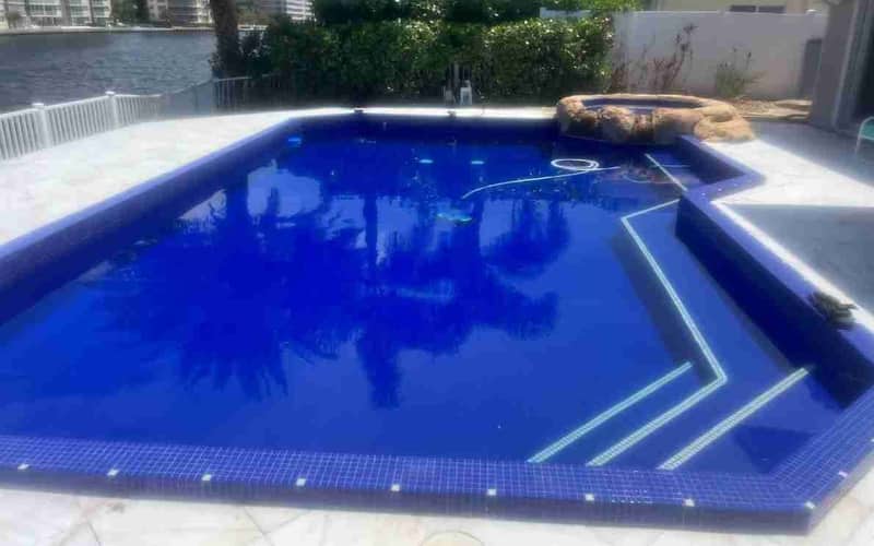 A deep blue tiled pool with a raised stone spa, white paving and a seawall with buildings beyond