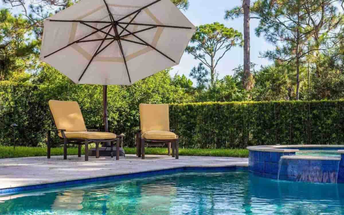 A pool with a stone spa spillway, two cushioned lounge chairs and an open umbrella beside a green hedge