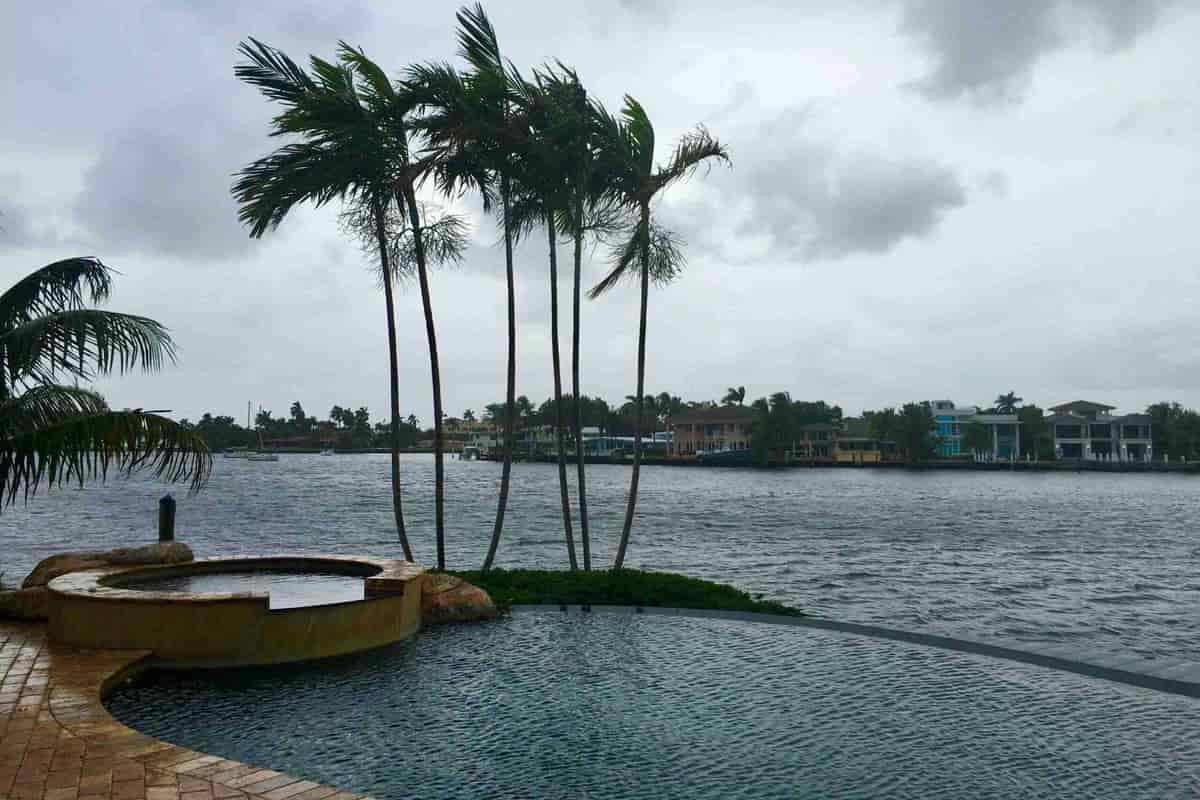 A raised round spa beside a pool edge that meets open water, with tall palm trees under a cloudy sky